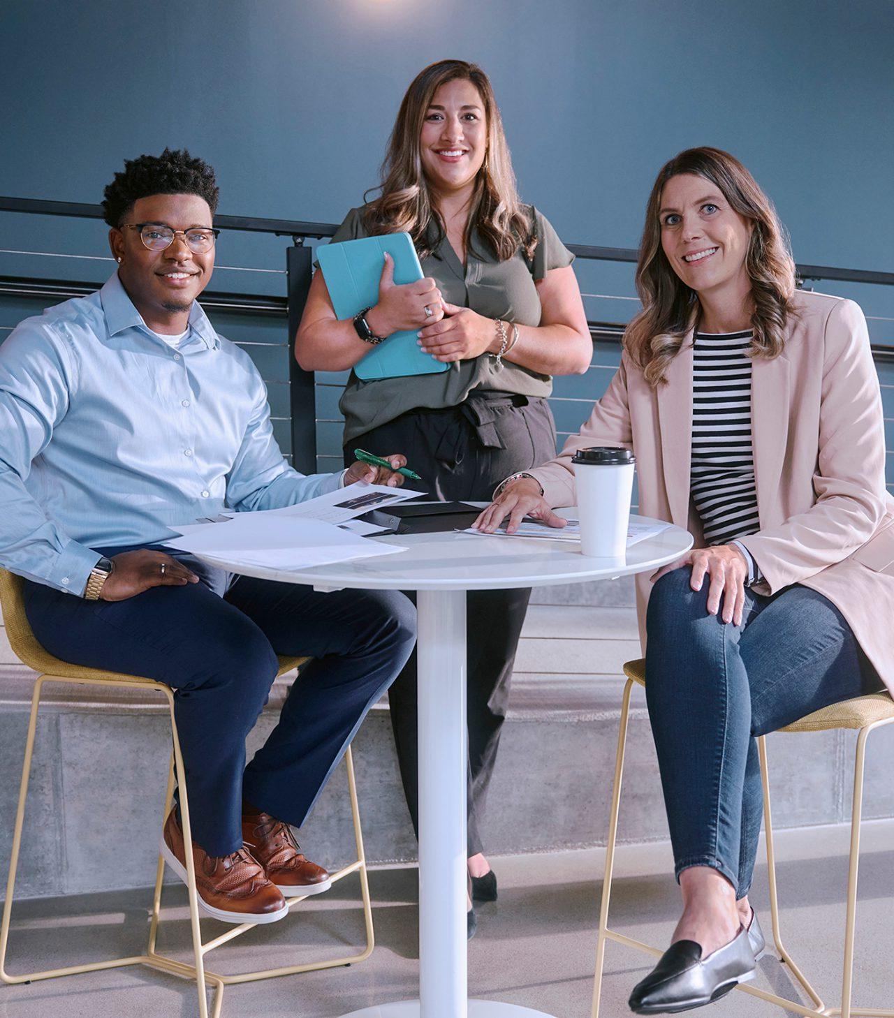 Two female employees and one male employee smiling and gathered around a tall white table.