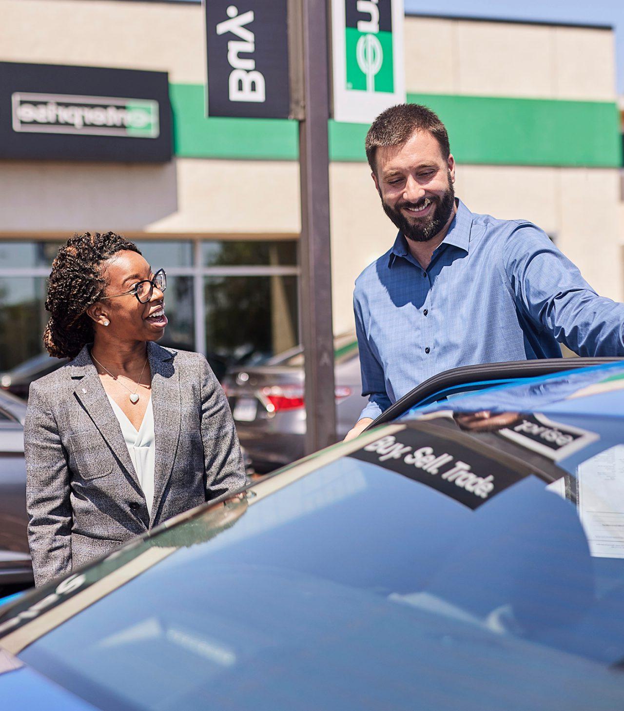 A gentleman, standing in front of a blue car, speaks with a lady at the Enterprise Car Sales lot.